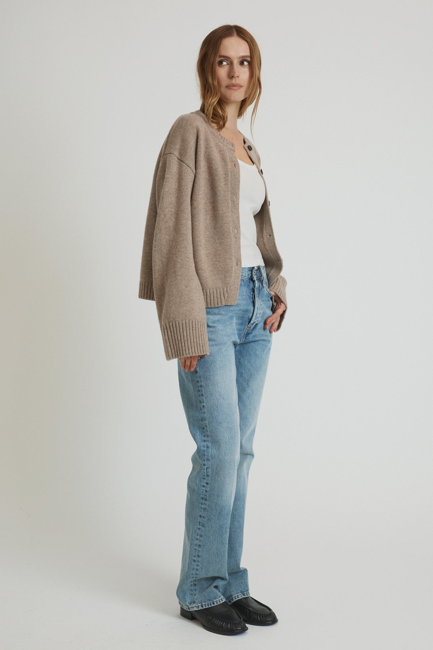 Woman wearing a beige cardigan, white top, blue jeans, and black shoes on a white background