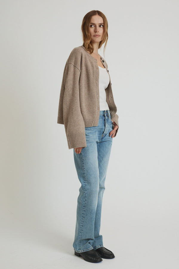 Woman wearing a beige cardigan, white top, blue jeans, and black shoes on a white background
